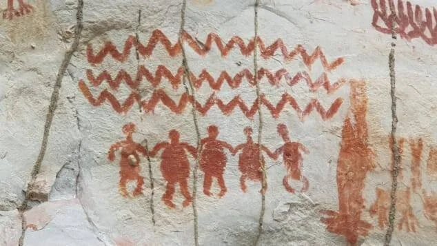 Pinturas rupestres en ocre rojo que muestran figuras humanas tomadas de la mano y líneas en zigzag sobre una pared de roca.