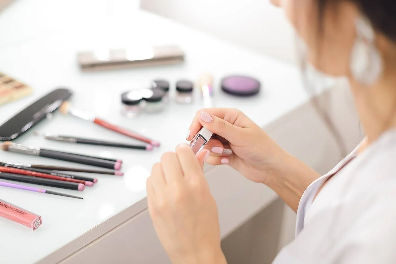 Mujer sostiene un labial mientras sobre la mesa hay lápices delineadores, sombras y otros cosméticos.