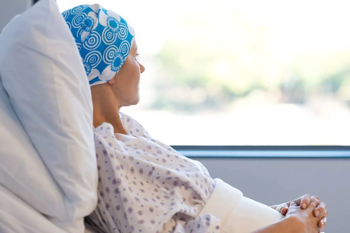 Mujer con pañuelo en la cabeza, posible paciente de cáncer, recostada en cama de hospital mirando por la ventana.