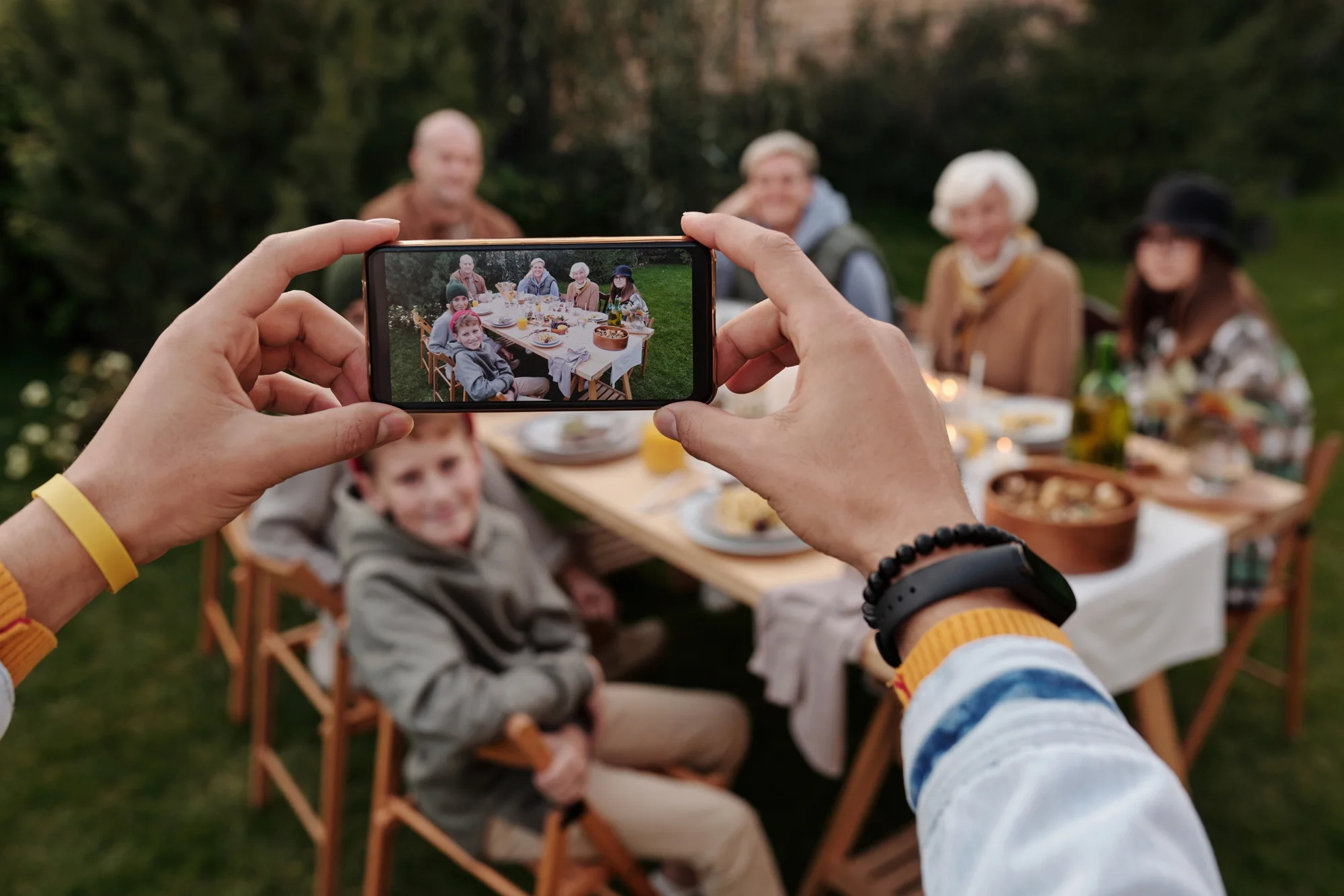 Manos sosteniendo un smartphone para fotografiar a un grupo familiar reunido en una mesa al aire libre.
