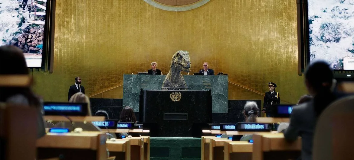 Un dinosaurio animado ocupa el podio de la Asamblea General de las Naciones Unidas, flanqueado por personas y el emblema de la ONU.