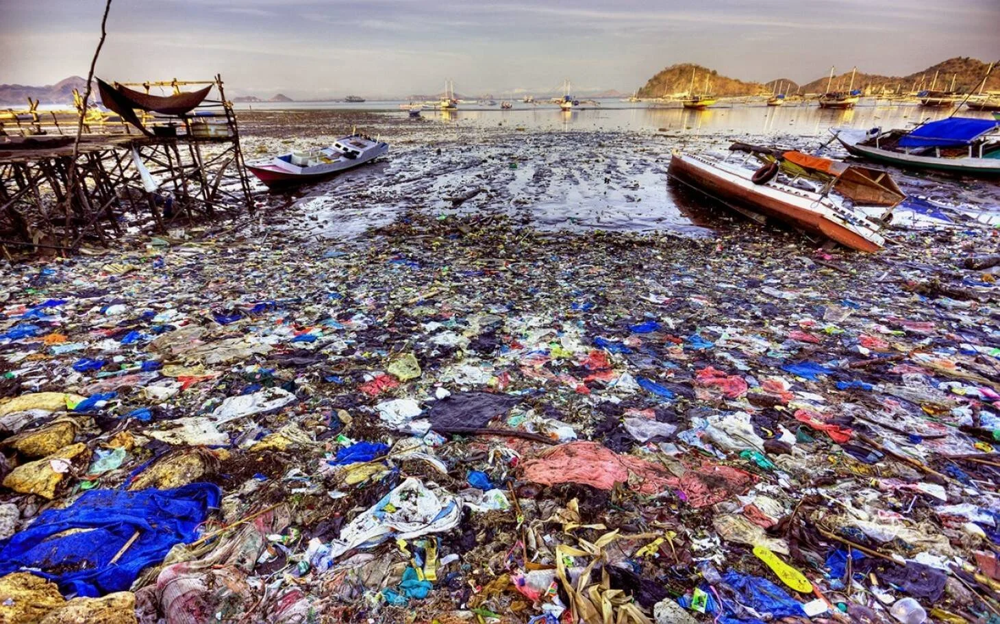 Bahía severamente contaminada con toneladas de residuos plásticos en la orilla y embarcaciones de pesca entre la basura flotante.