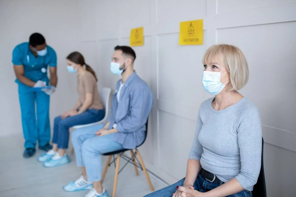 Tres personas con mascarilla sentadas en sillas de una sala de vacunación mientras un profesional sanitario atiende a una de ellas.