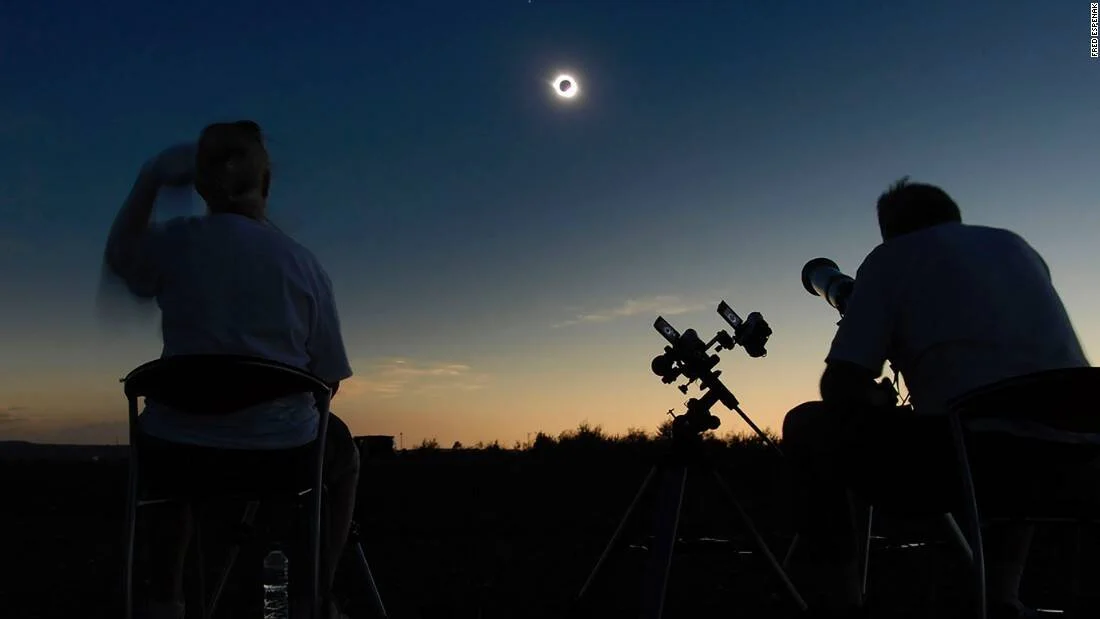 Dos personas observan y fotografían un eclipse solar total con telescopios al atardecer, con la corona solar visible en el cielo.