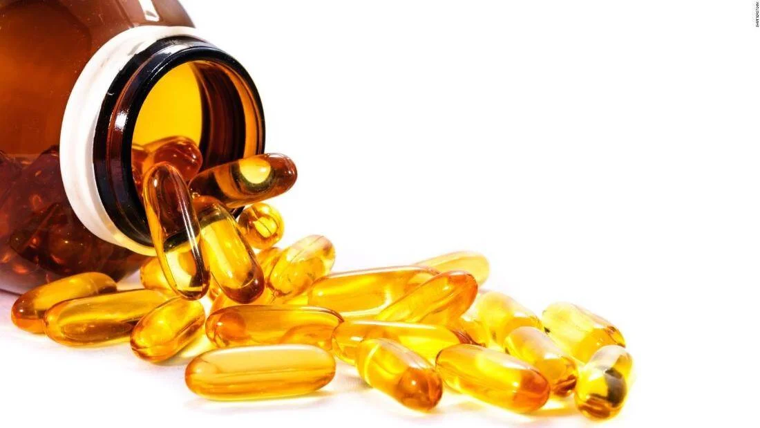Frasco de suplementos volcado con cápsulas de gel de aceite de pescado color ámbar esparcidas sobre fondo blanco.