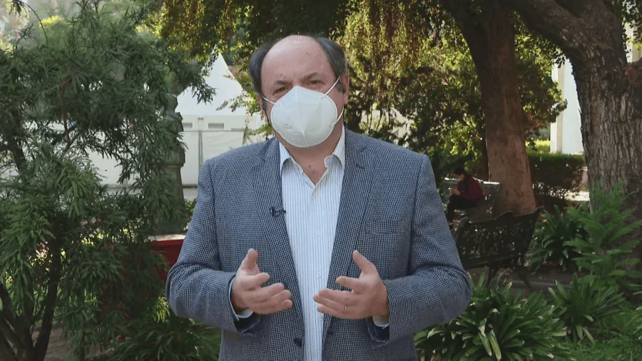 Hombre con saco gris y cubrebocas blanco gesticula mientras habla frente a cámaras en un parque arbolado.