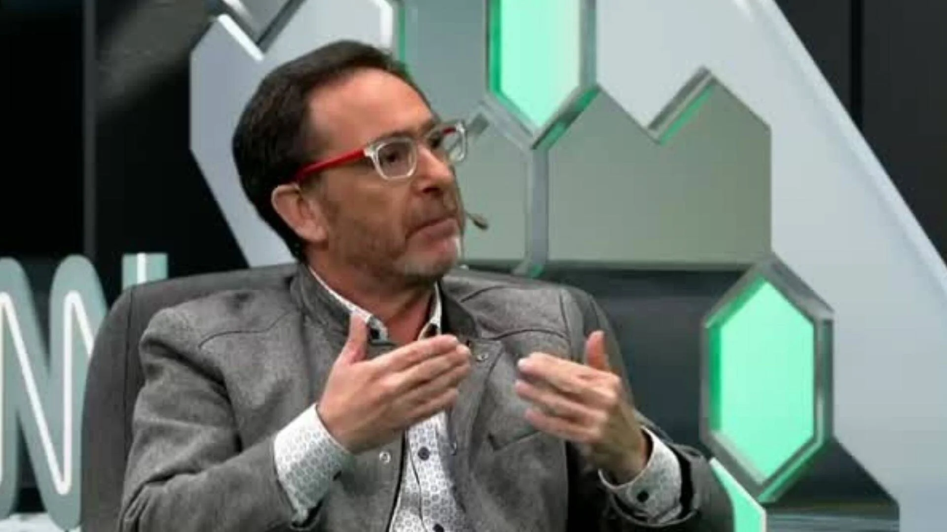 Hombre con anteojos de marco rojo gesticula con las manos mientras habla en un estudio de televisión con fondo de hexágonos verdes.
