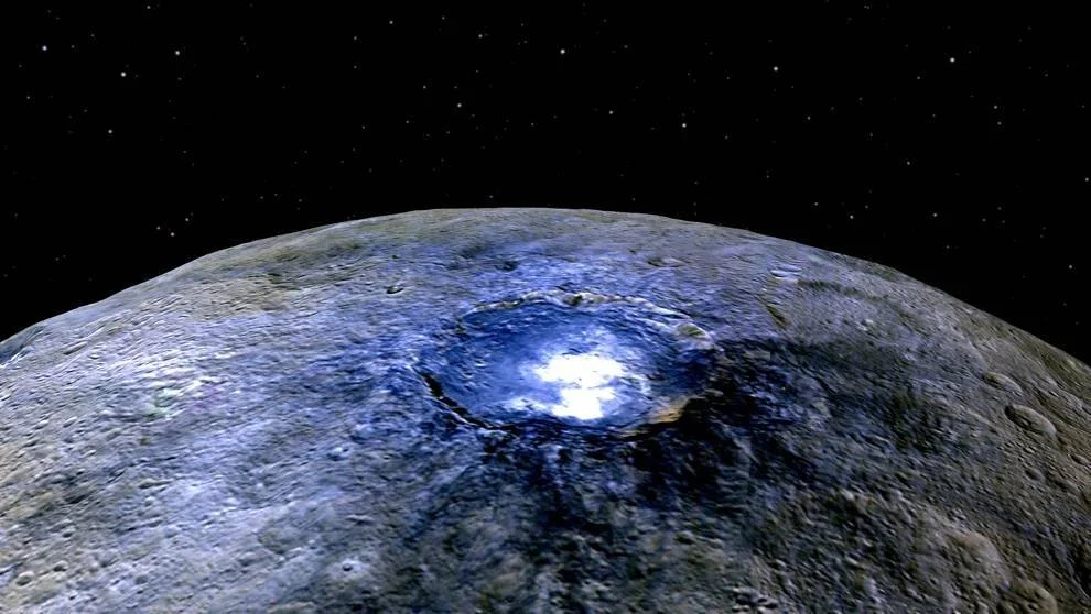 Vista orbital de un gran cráter con depósitos brillantes blancos y azulados sobre la superficie gris de un cuerpo celeste, con estrellas al fondo.