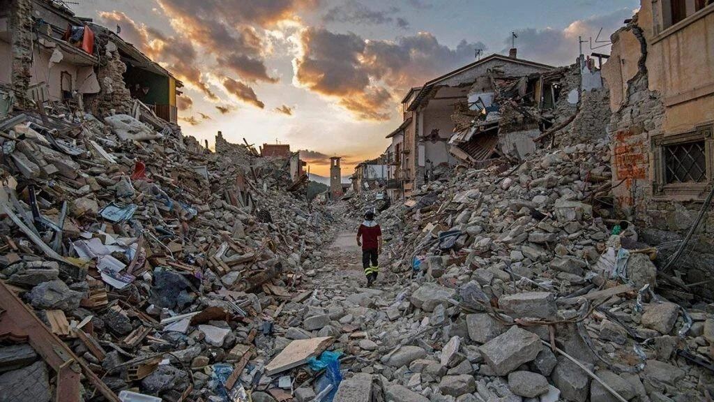 Una persona camina sola por una calle completamente destruida, rodeada de escombros de edificios derrumbados al atardecer.