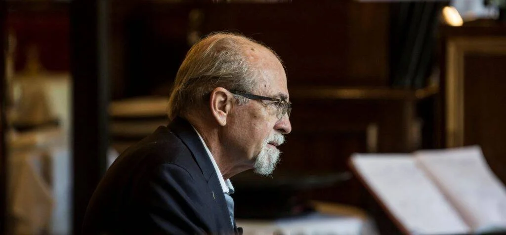 Hombre mayor de cabello cano y barba blanca, con anteojos y traje oscuro, sentado frente a un atril con partituras en una sala con estanterías.