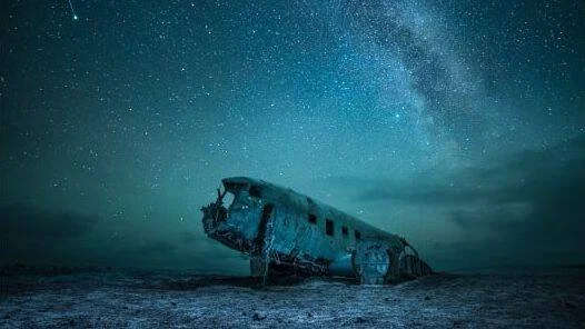 Restos oxidados de un avión abandonado en un paisaje árido bajo un cielo nocturno con la Vía Láctea visible.