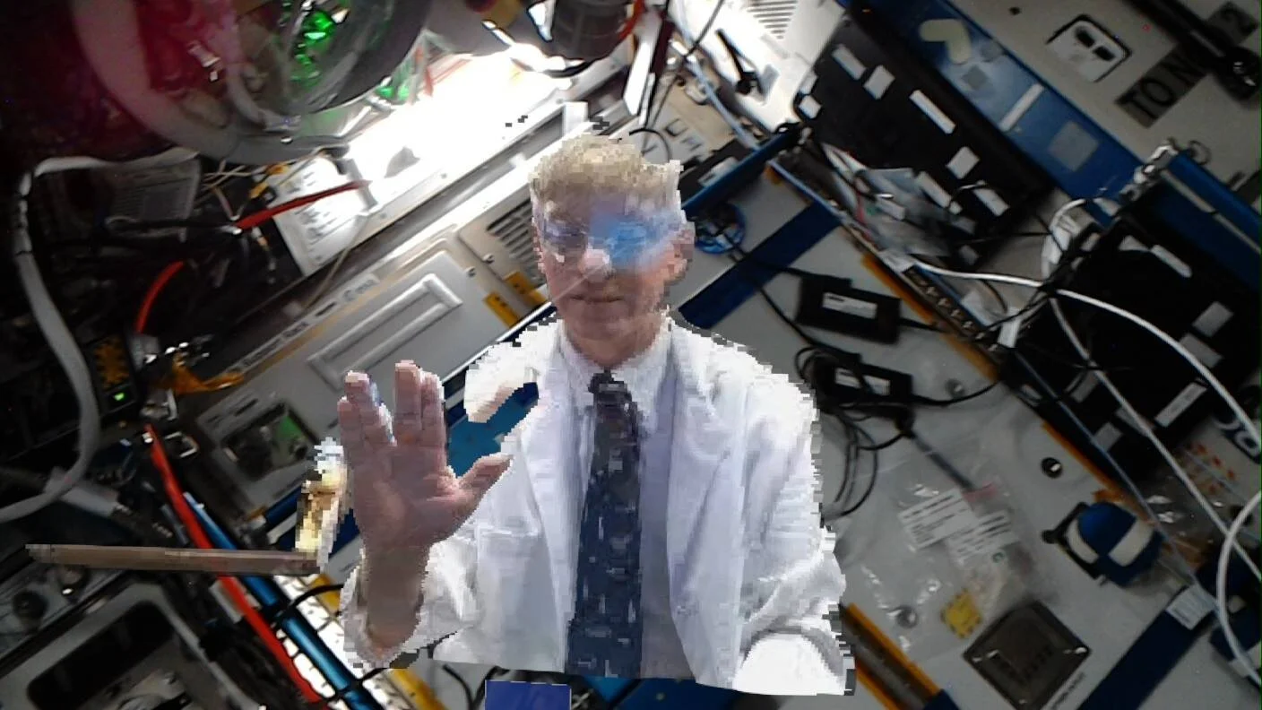 Holograma pixelado de un hombre con bata blanca y corbata, proyectado en el interior de una estación espacial rodeada de equipos y cables.