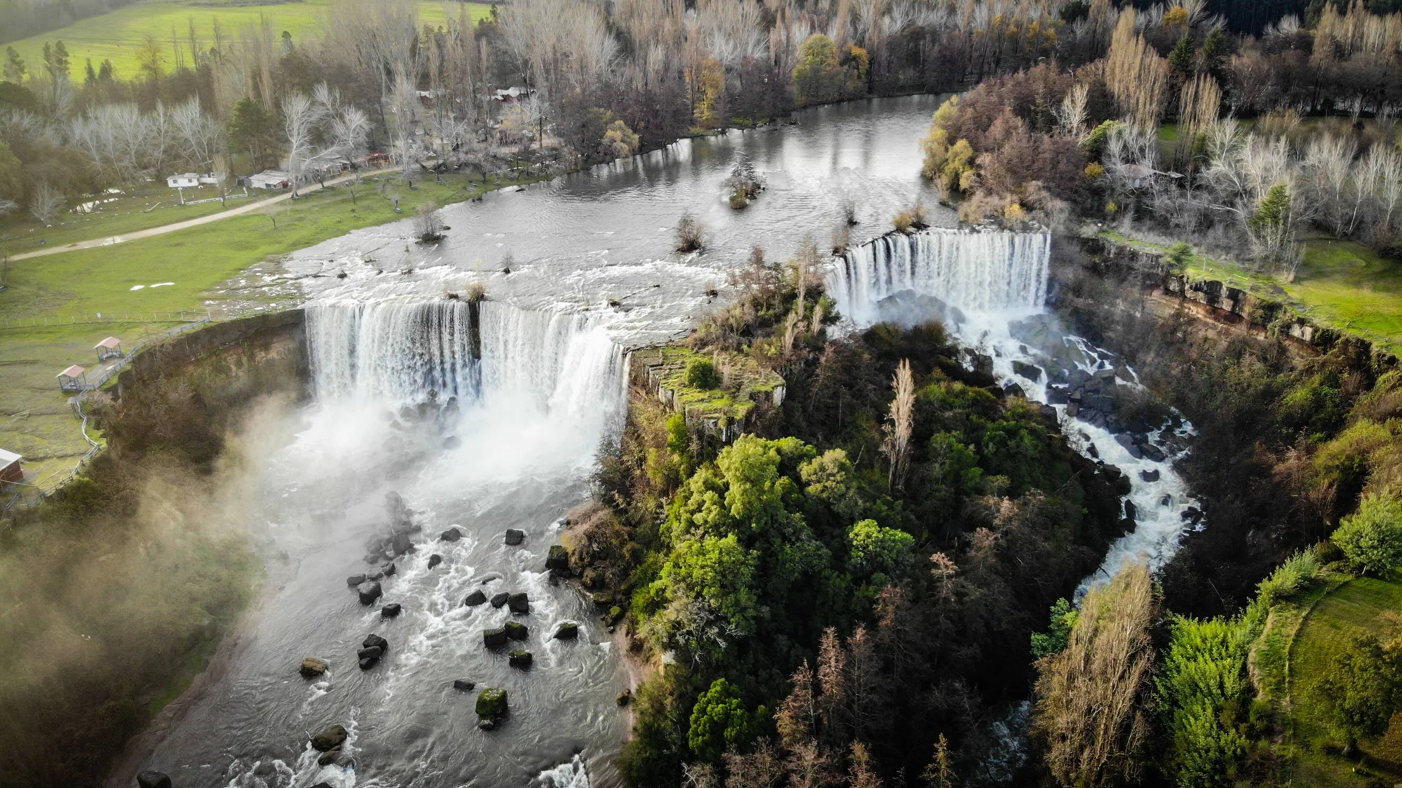 Panorámica aérea de un conjunto de cascadas que caen sobre rocas basálticas rodeadas de bosque nativo y praderas verdes.