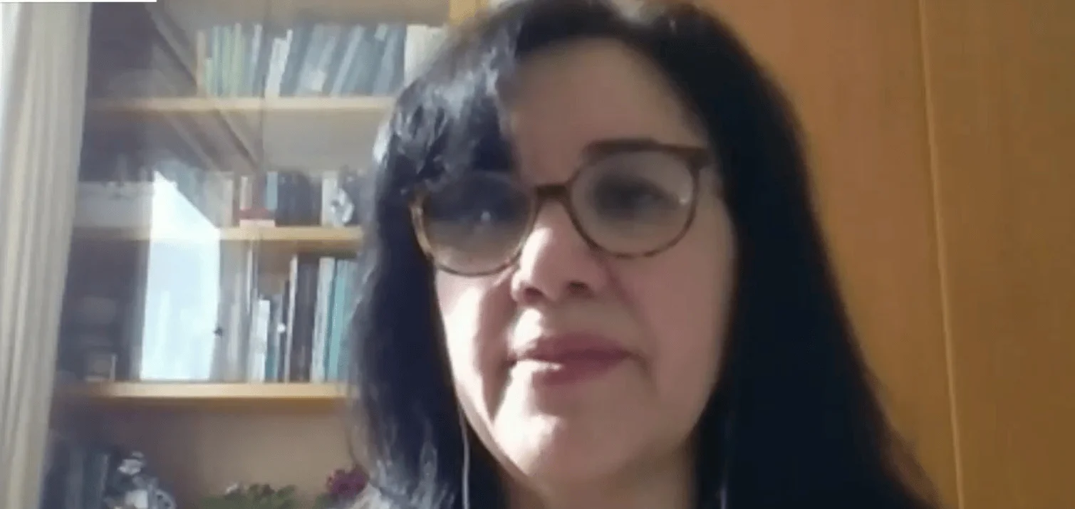 Mujer de cabello oscuro y gafas habla durante una videollamada con una estantería de libros al fondo.