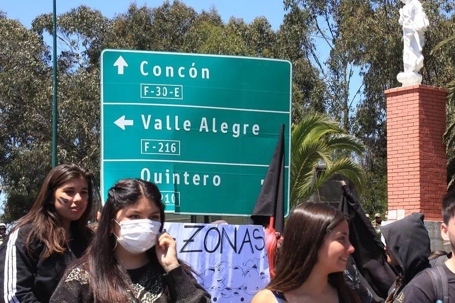 Manifestantes, una con mascarilla y cartel que dice 'Zonas', protestan frente a señal vial hacia Concón, Valle Alegre y Quintero.