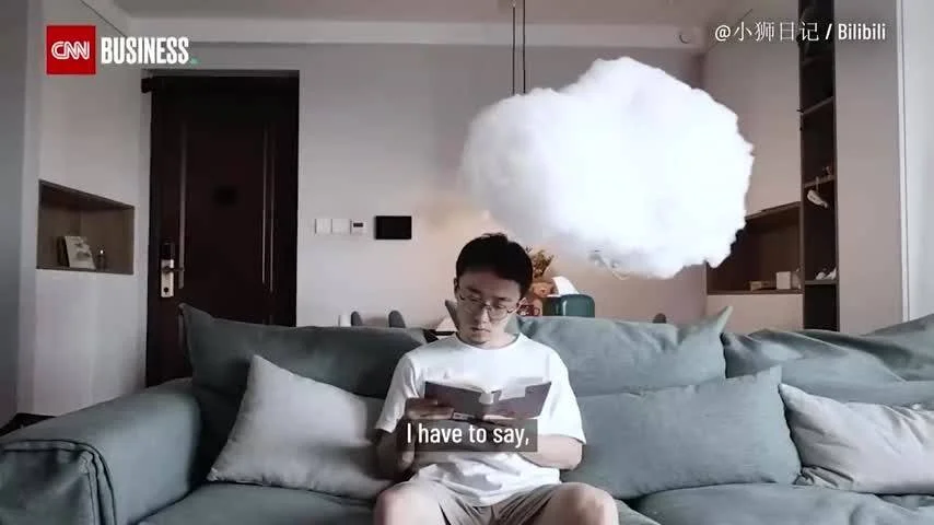 Hombre con gafas lee un libro en un sofá mientras una nube artificial flota sobre su cabeza, en un video de Bilibili para CNN Business.