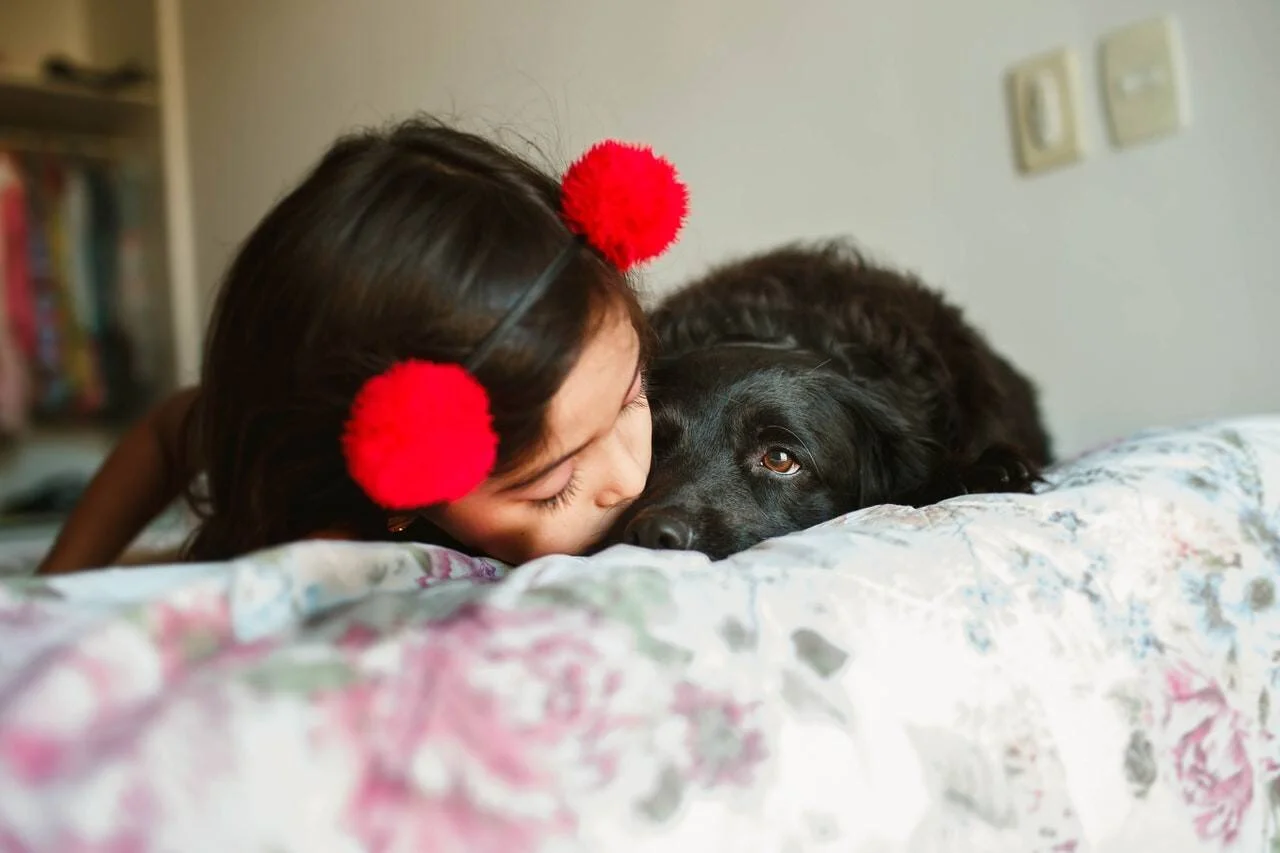 Una niña con pompones rojos en el cabello da un beso cariñoso a un perro negro acostado en una cama.