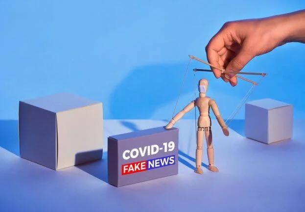 Una mano manipula una marioneta de madera con mascarilla junto a una caja con la leyenda 'COVID-19 Fake News', simbolizando la desinformación.