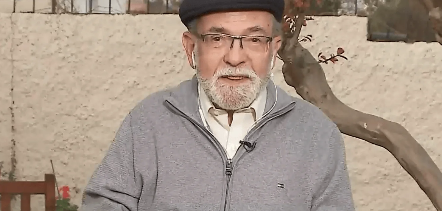 Hombre mayor con boina negra, gafas y barba blanca habla con micrófono de solapa frente a una pared exterior.