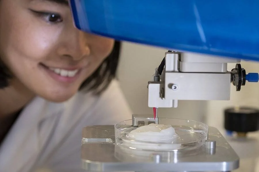 Investigadora con bata observa de cerca una bioimpresora 3D depositando material sobre una muestra en una placa de Petri.