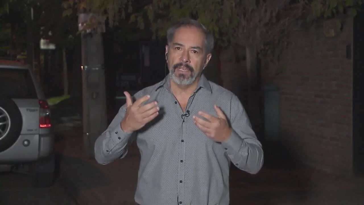 Hombre de cabello entrecano y barba gris habla a cámara con micrófono de solapa en exteriores nocturnos.