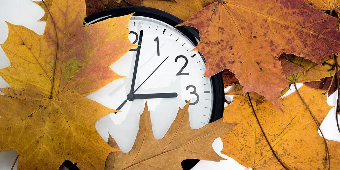 Reloj analógico blanco rodeado de hojas secas de otoño en tonos naranja y marrón, evocando el cambio de hora estacional.