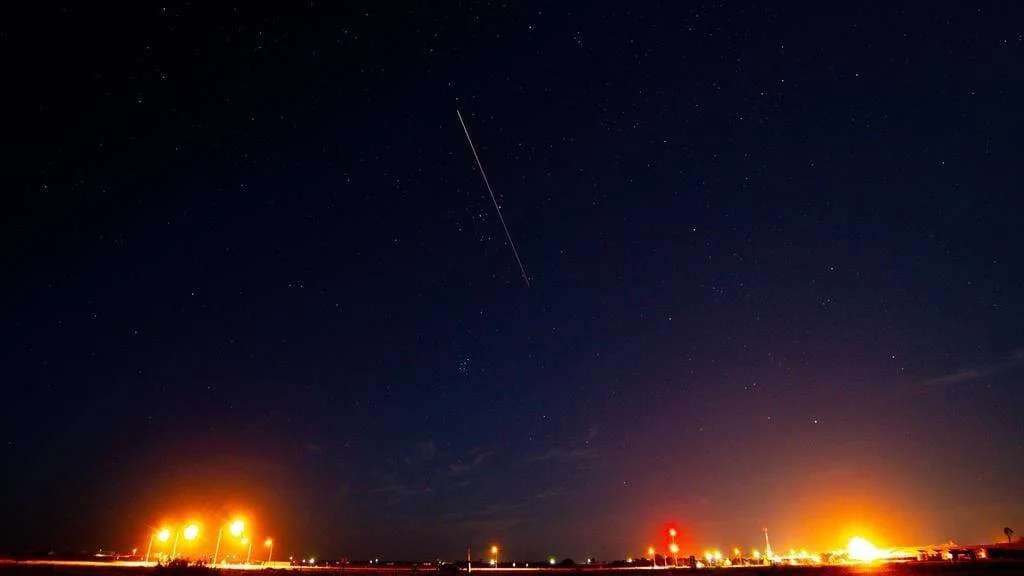 Rastro de luz de un cohete atraviesa el cielo nocturno estrellado sobre una zona iluminada en tierra.