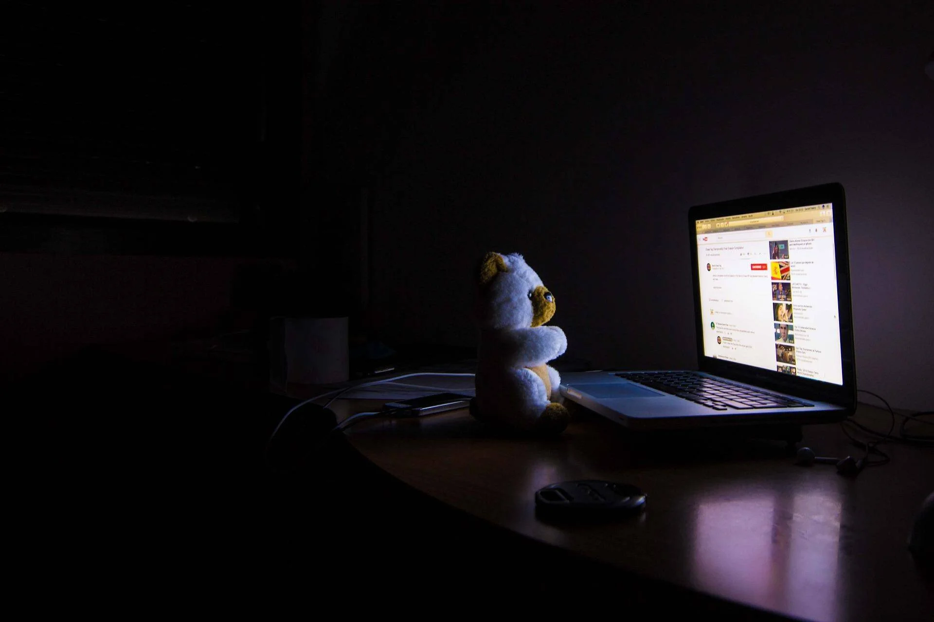 Osito de peluche sentado ante una laptop con YouTube abierto en una habitación a oscuras, iluminado solo por la pantalla.
