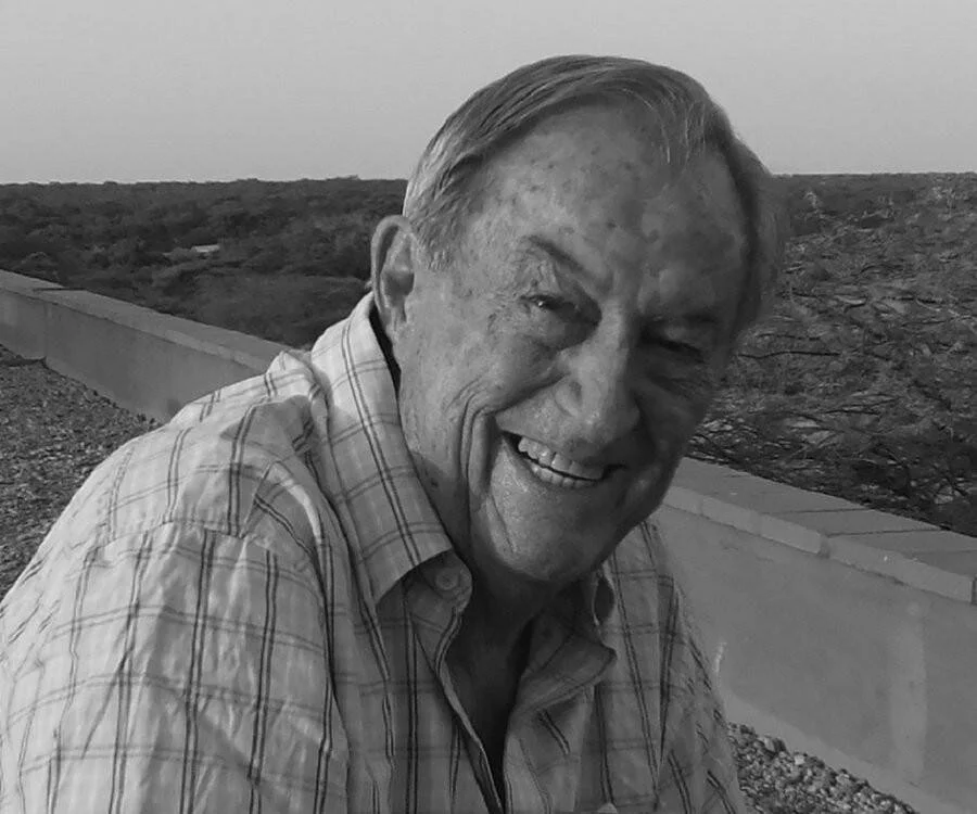 Hombre mayor sonriente con camisa a cuadros, fotografiado en exteriores con vegetación de fondo, en blanco y negro.