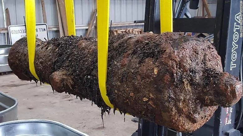 Gran bomba sin explotar, severamente oxidada y cubierta de sedimento, suspendida con correas amarillas desde un montacargas Toyota.