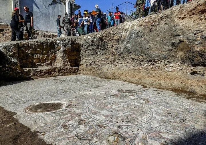 Grupo de personas observa desde arriba un gran mosaico romano con motivos circulares recién excavado en un sitio arqueológico.
