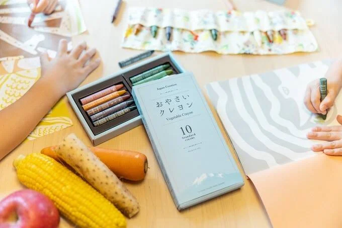 Caja de crayones vegetales japoneses 'Vegetable Crayon' de 10 colores junto a maíz, zanahoria y manzana, mientras niños dibujan al fondo.