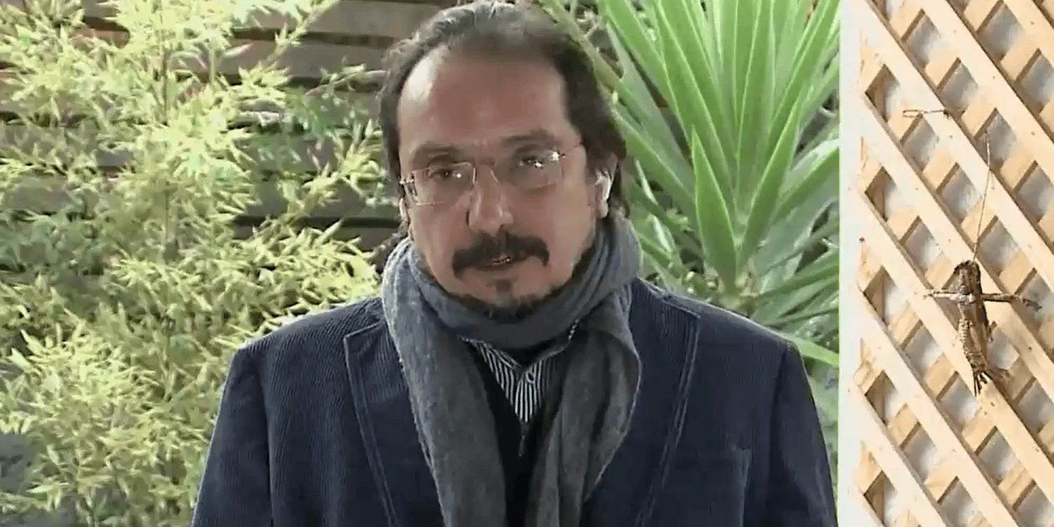 Hombre de mediana edad con anteojos, bufanda gris y saco azul frente a plantas verdes en un jardín.