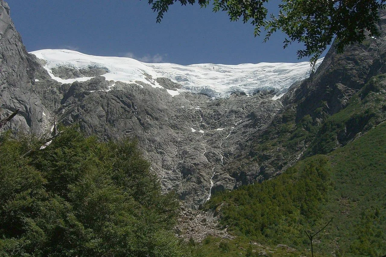 Glaciar sobre una empinada pared rocosa de montaña, con deshielo que desciende por la ladera y vegetación andina en primer plano.
