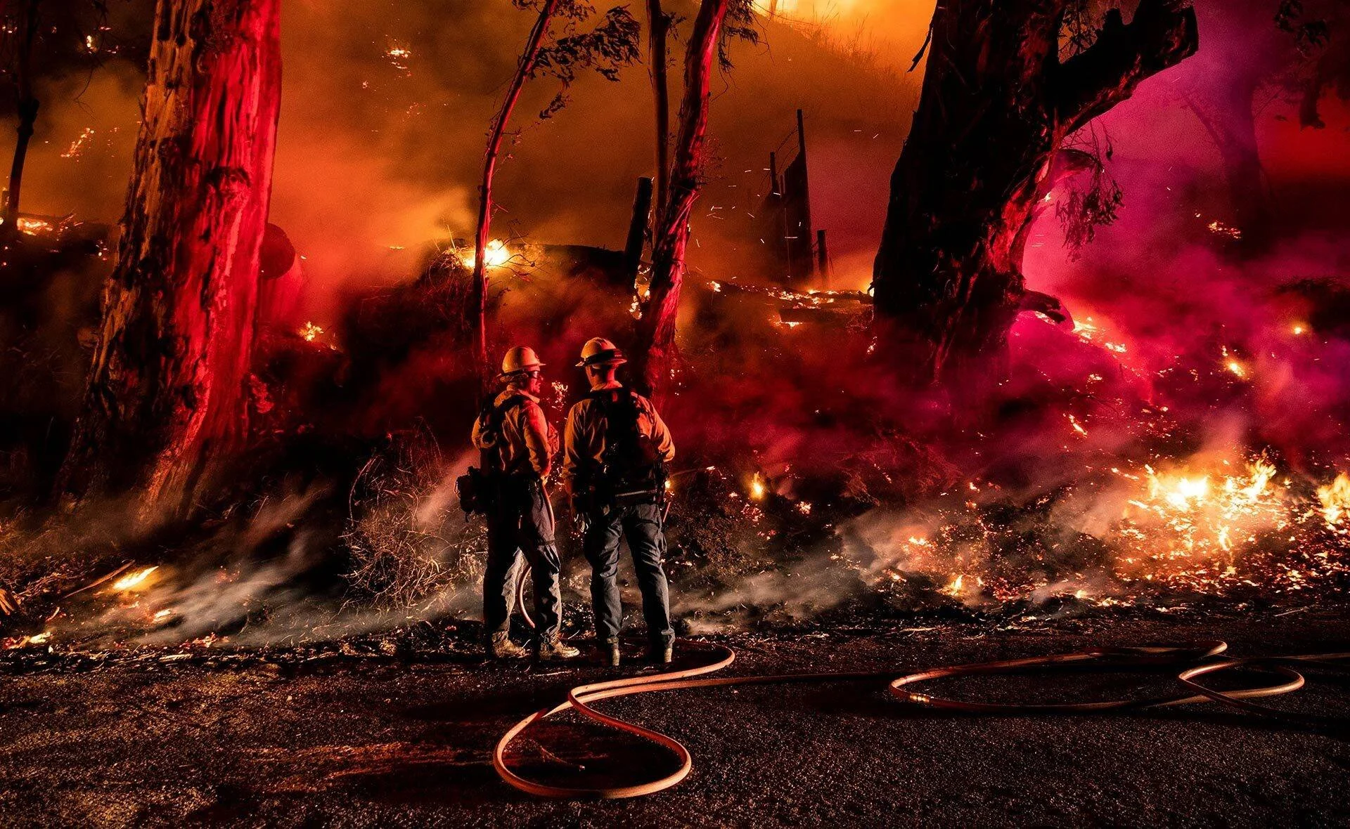 Dos bomberos con cascos observan un violento incendio forestal nocturno rodeados de llamas, humo y árboles en llamas.