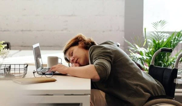 Mujer en silla de ruedas recostada sobre un escritorio con laptop, aparentemente dormida o agotada en su lugar de trabajo.