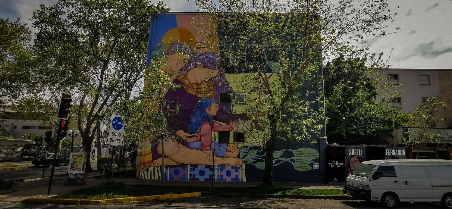 Gran mural callejero con dos figuras humanas entrelazadas en colores vivos cubre la fachada de un edificio en una avenida arbolada.