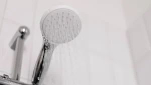 Cabezal de ducha blanco y cromado con agua fluyendo sobre fondo de azulejos blancos.