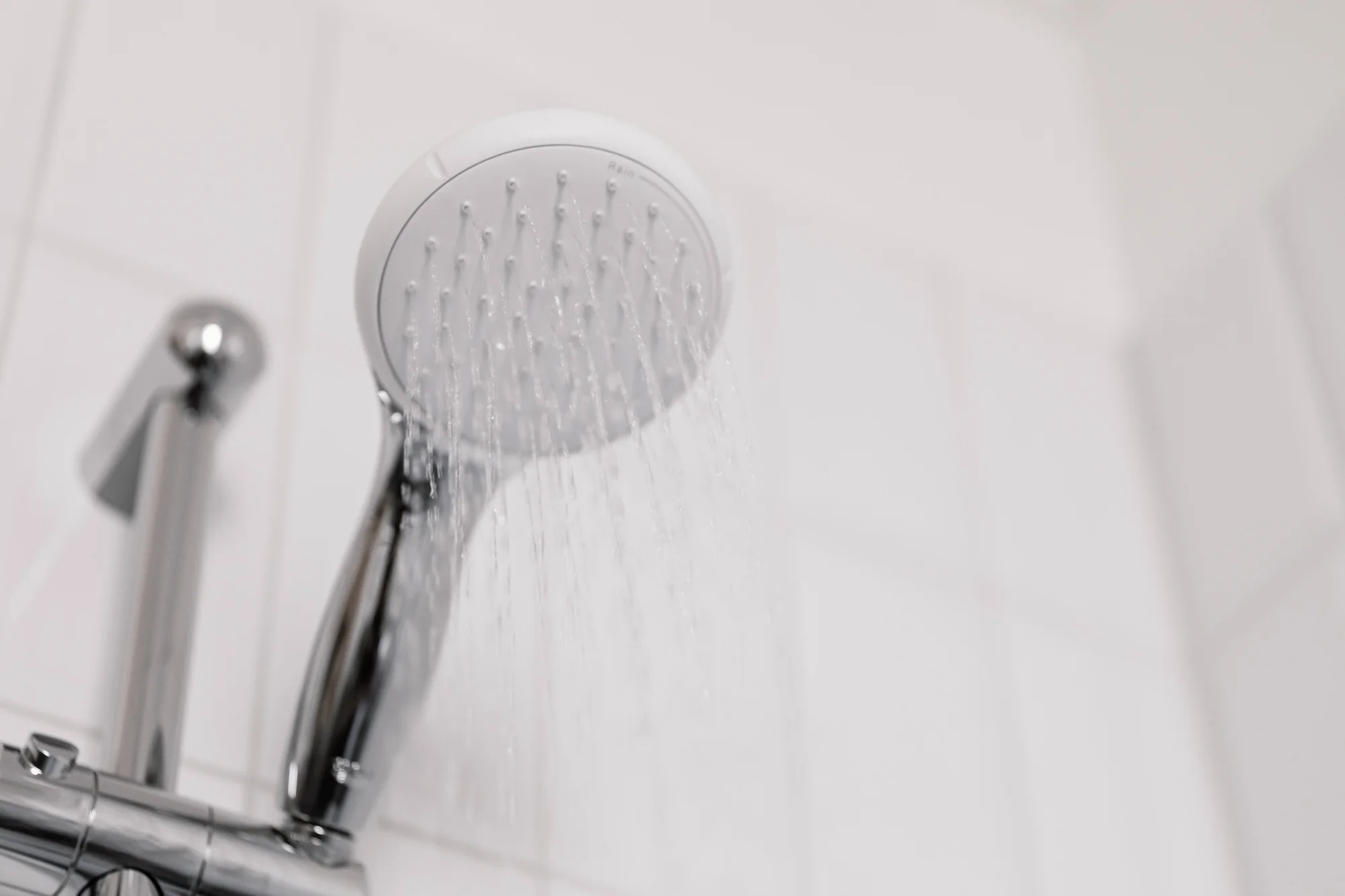 Cabezal de ducha blanco y cromado con agua fluyendo sobre fondo de azulejos blancos.