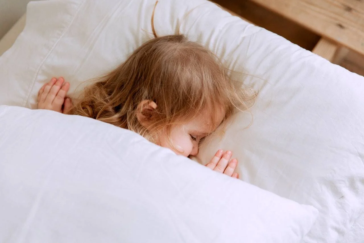 Niña de cabello castaño claro durmiendo acurrucada entre almohadas y sábanas blancas.