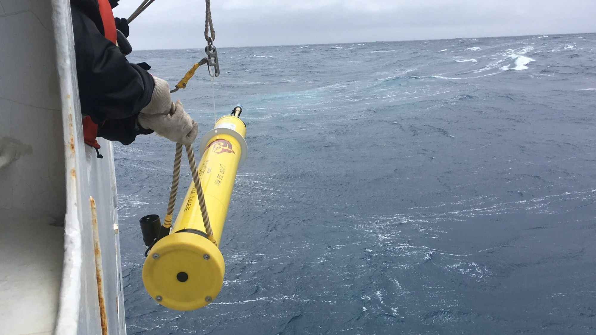 Investigador sostiene una boya oceanográfica cilíndrica amarilla mientras la despliega al mar agitado desde la borda de un barco.