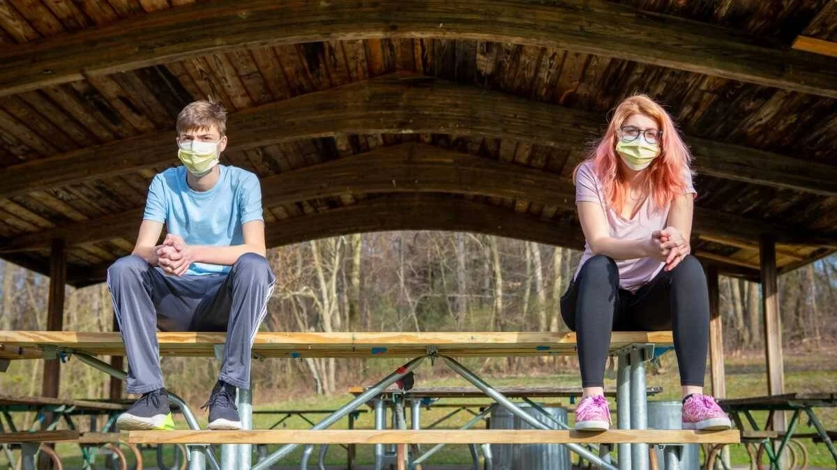 Dos adolescentes con mascarillas sanitarias sentados con distancia social sobre una mesa de picnic bajo un cobertizo de madera.
