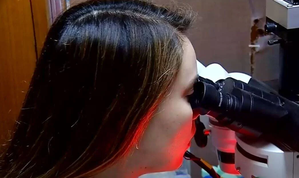 Mujer joven mirando a través del ocular de un microscopio iluminado con luz roja en un laboratorio.
