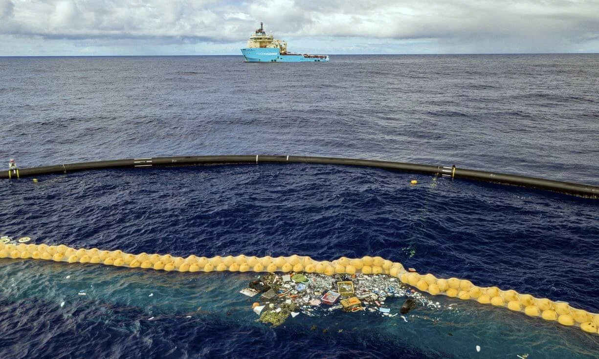 Sistema de barrera flotante con boyas captura residuos plásticos en el océano, con un barco de limpieza al fondo.