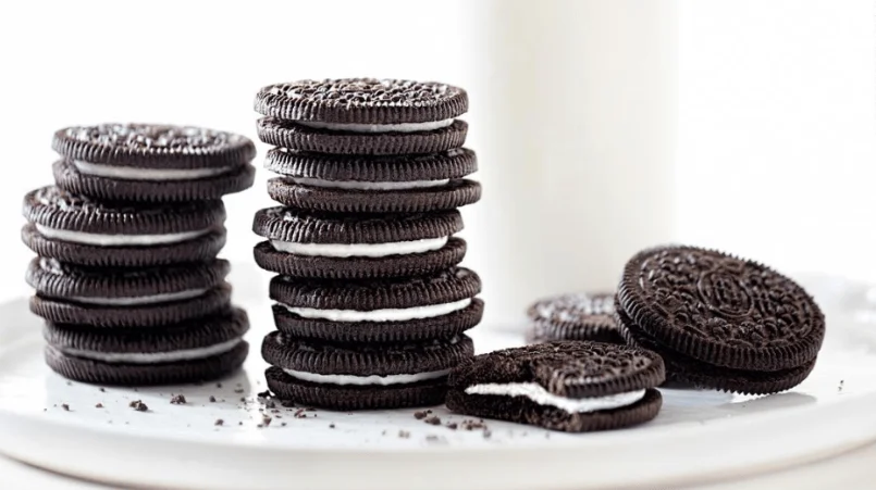 Dos pilas de galletitas de chocolate rellenas con crema blanca sobre un plato, con una galletita abierta al costado y un vaso de leche al fondo.