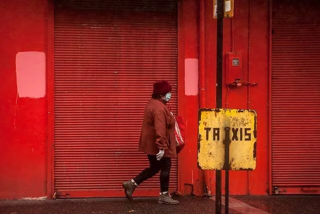 Una persona con barbijo y abrigo rojo camina frente a una persiana metálica roja junto a un cartel oxidado de taxis.