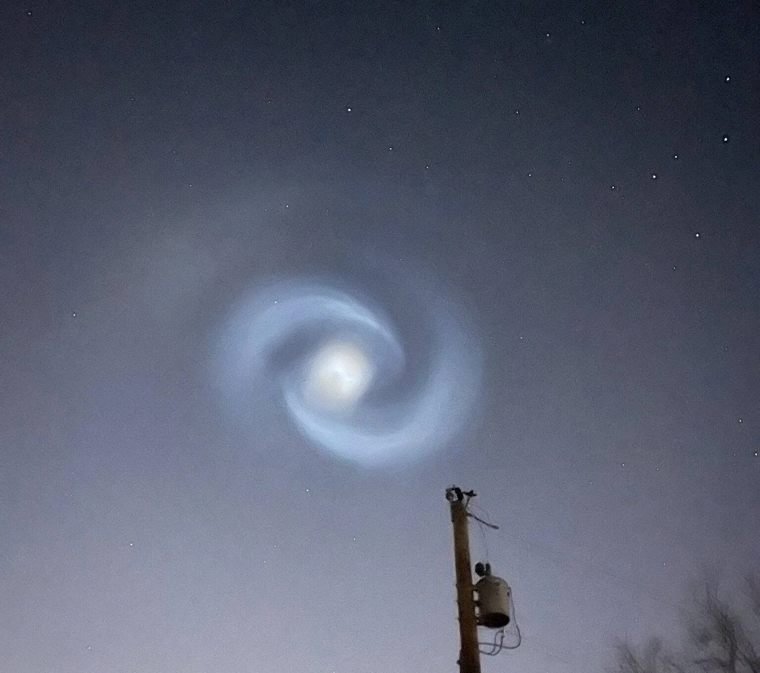 Espiral de luz azulada con núcleo brillante gira en el cielo nocturno sobre un poste eléctrico, posiblemente generada por combustible de cohete.