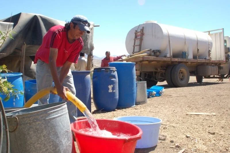 Un hombre llena baldes y tinas con agua proveniente de un camión cisterna en zona árida sin acceso a red de agua potable.