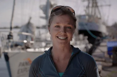 Mujer sonriente con gafas de sol en la cabeza frente a embarcaciones en un puerto deportivo.