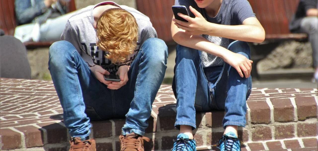 Dos jóvenes sentados en un escalón de ladrillos mirando sus teléfonos móviles al sol.